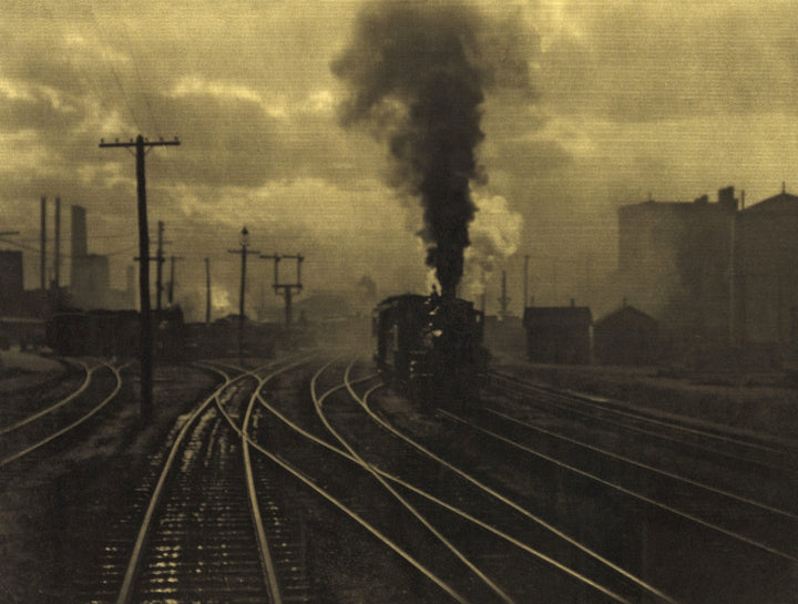 A steam locomotive with billowing smoke travels along curved railway tracks through an industrial urban landscape under hazy skies.