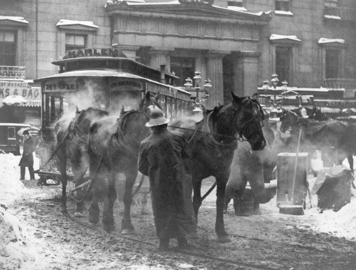 Horse-drawn streetcar with passengers in snowy urban street lined with buildings, photographed in black and white.