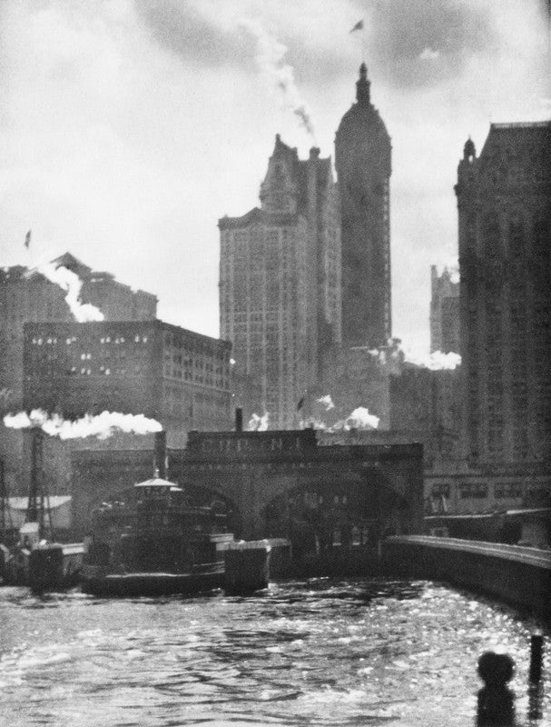 Black and white photograph of New York City skyline with tall buildings, a bridge, and steamboat on water below.