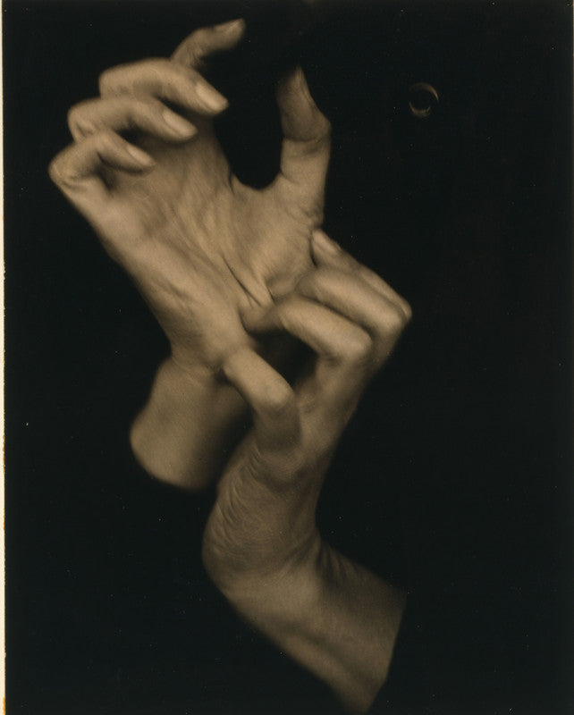 Two pale hands intertwined against a dark background in a sepia-toned photograph.