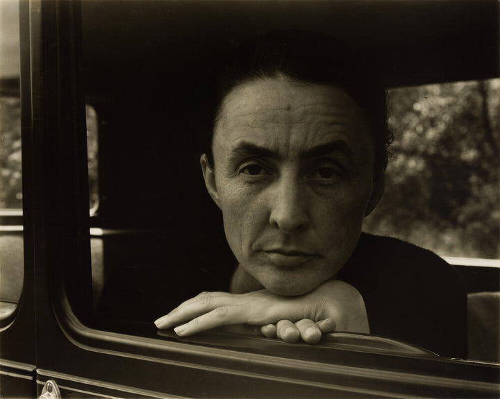 A man with a stern expression leans toward a car window, his hand resting on the frame, in a black and white photograph.