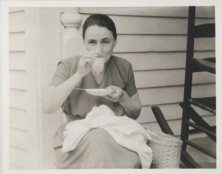 Black and white photograph of a woman seated on a porch, drinking from a cup while holding a plate.