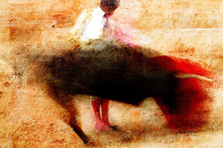 A matador in white clothing holds a red cape while a dark bull charges across a sandy, textured background in warm earth tones.