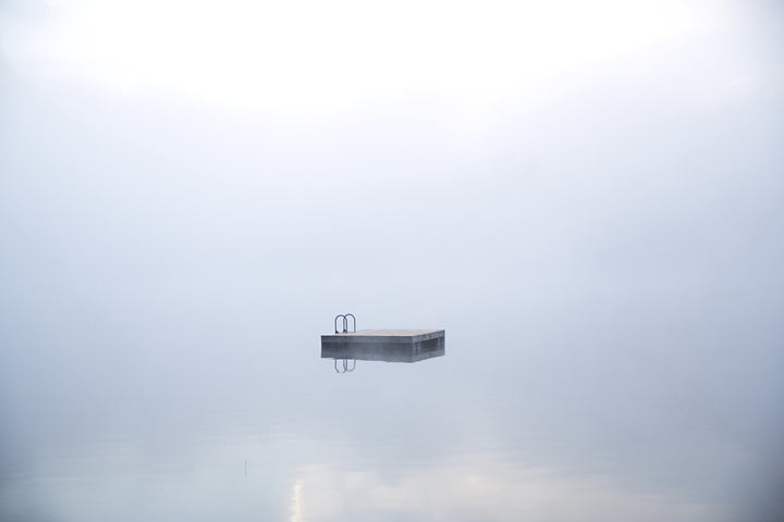 A concrete platform with metal railings floats in a misty, gray sky above calm water.