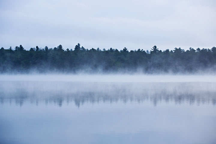 Misty morning landscape with forested shoreline reflected in calm blue water beneath overcast sky.
