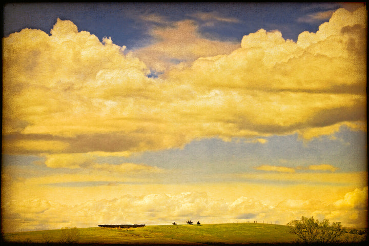 Golden prairie landscape with distant cattle roundup under towering yellow clouds and blue sky.