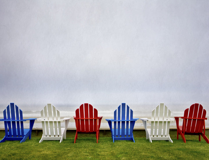 Six colorful Adirondack chairs in blue, white, and red arranged in a row on grass facing the sea.
