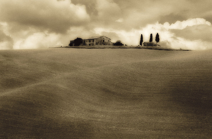A sepia-toned photograph shows an isolated farmhouse with cypress trees in a flat, plowed field under cloudy sky.