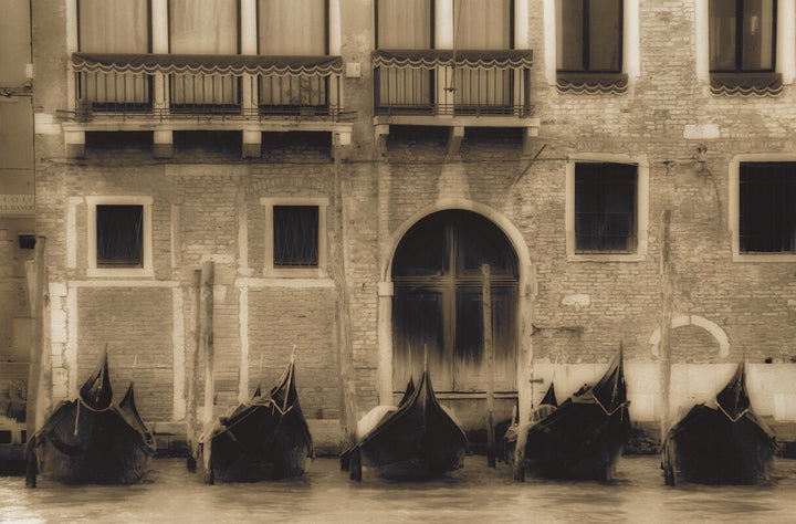 Five black gondolas moored in front of a cream-colored Venetian building with arched doorway and balconies.