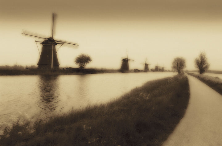 Sepia-toned landscape featuring silhouetted windmills reflected in still water with bare trees and a canal.