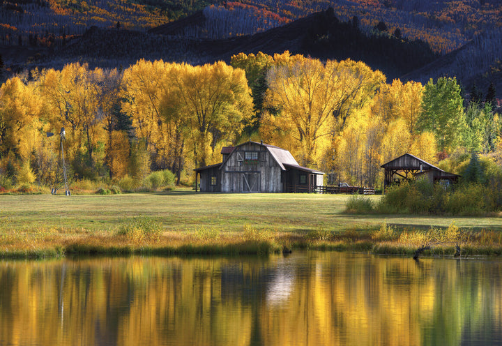 A rustic barn and outbuildings stand in a meadow beside a reflective pond, framed by golden aspen trees and forested mountains.