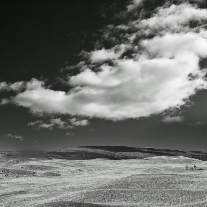Black and white photograph depicting a vast plowed field beneath a dramatic cloudy sky with a distant hill.