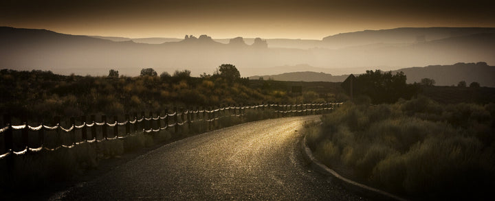 A long straight road with fence line stretches toward misty mountains under golden-brown atmospheric haze.