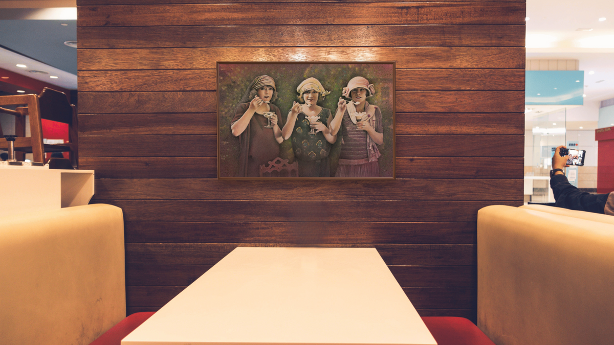 Ice Cream Ladies by Kirsten Söderlind, shown at 28 × 42 in (71.1 × 106.7 cm) on canvas in Arber Warm Walnut, restaurant